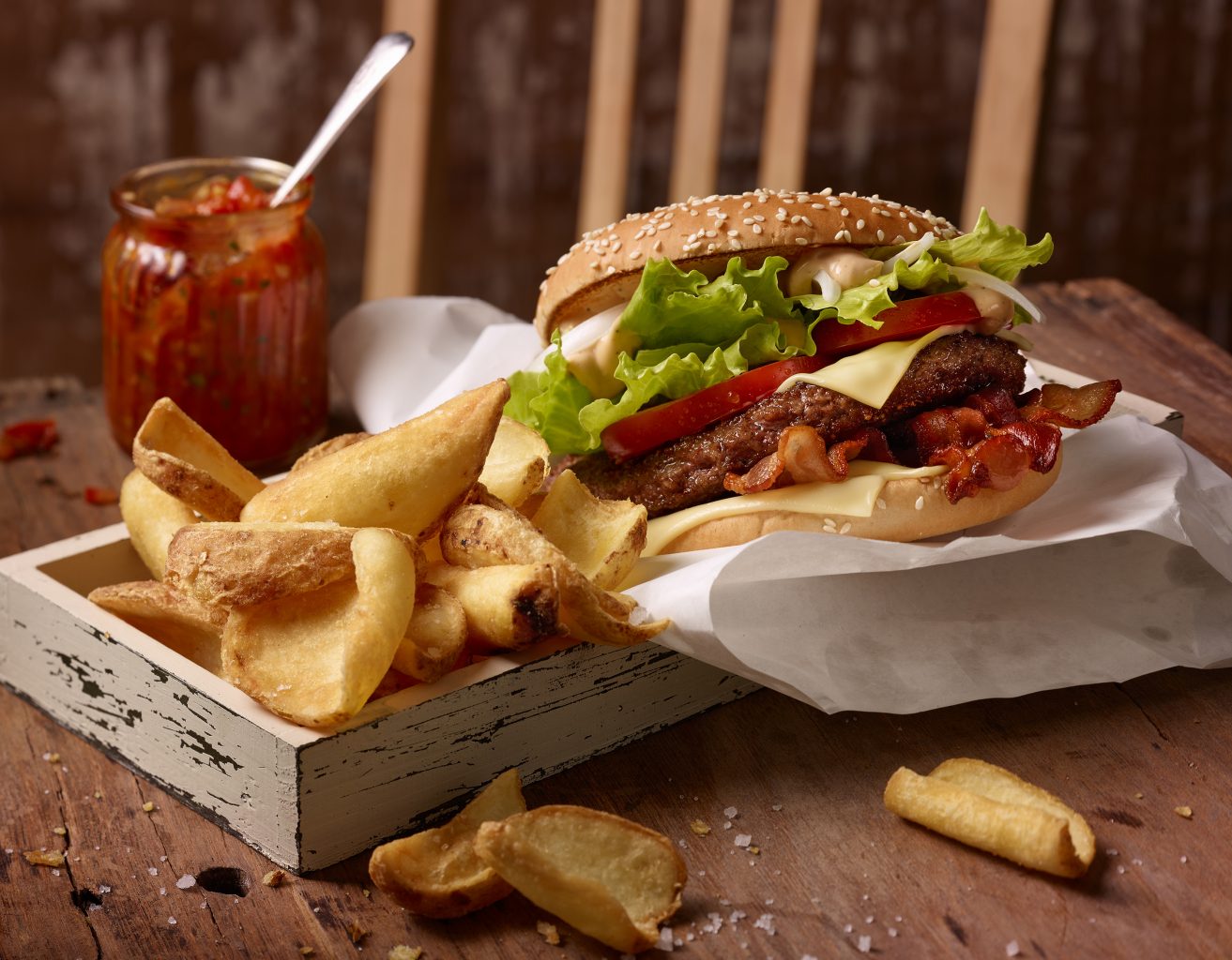 fast food fotografie von big tasty bacon und curly fries pommes auf alten holztablett, stuhl steht im Hintergeund, sosse ist mit Löffel im Glasgefäß