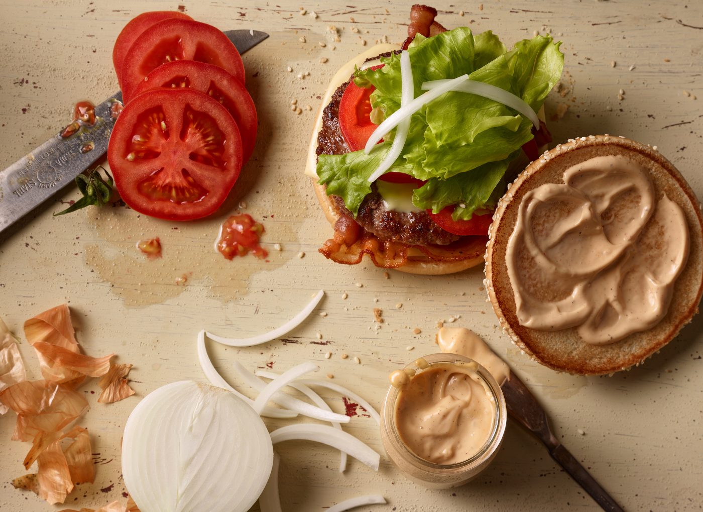 fast food fotografie zusammenbau eines cheeseburgers aus buns sosse zwiebeln tomatenscheiben salat messer und sossenschale stehen daneben lebendige situation mit gekruemmel und gekleckse vogelperspektive