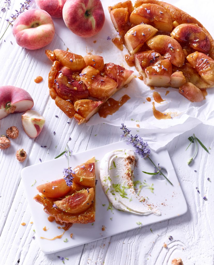 angechnittener Pfirsich Kuchen liegt auf weiss gestichenem Holzbrettern aussenrum liegen blumenzweige obst und ander props