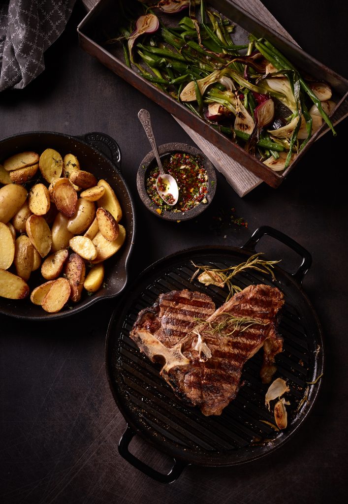 Dry Aged T-Bone Steak gegrillt in der Grillpfanne liegend mit Spargel Kartoffeln Gremolata Chimichurri Salsa auf dunklem Metall Untergrund mit Salsa Knoblauch Pfeffer hochformat dark food photography münchen
