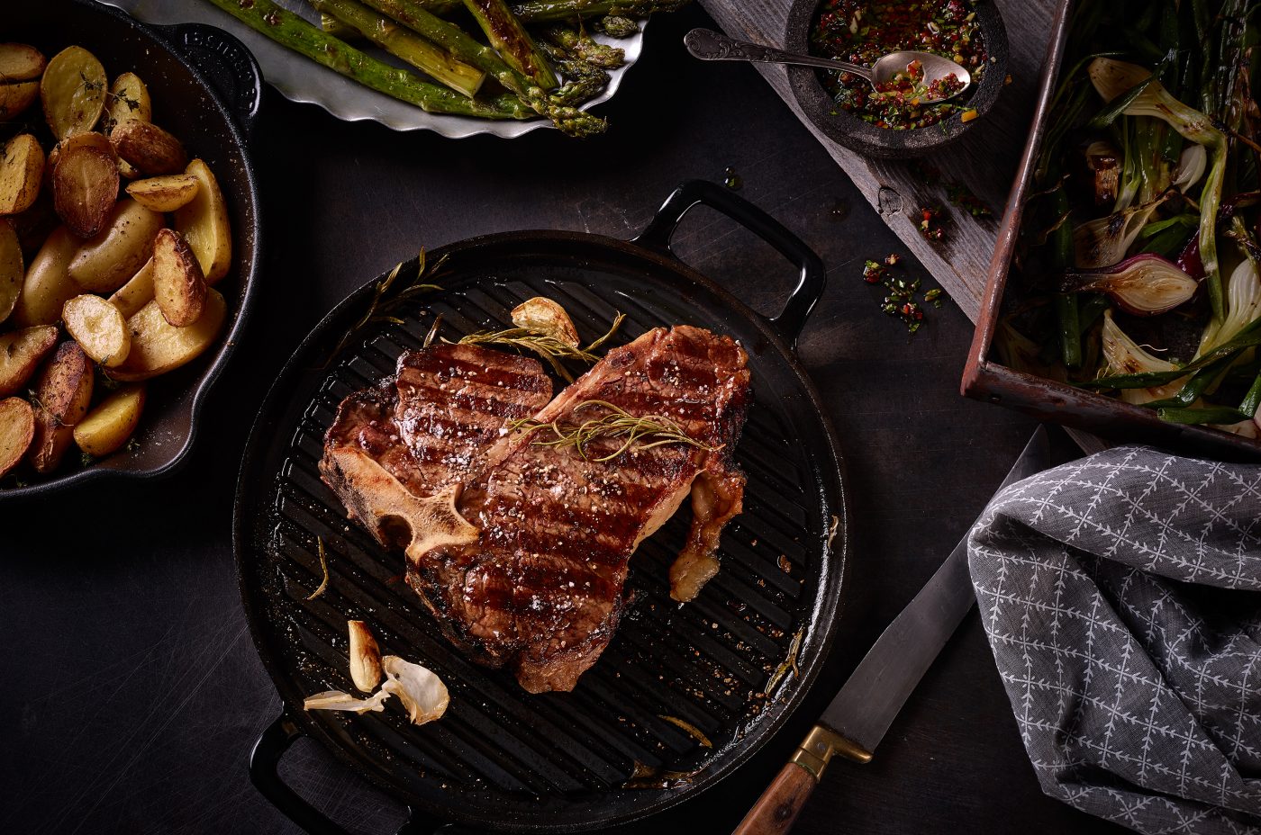 Dry Aged T-Bone Steak gegrillt in der Grillpfanne liegend mit Spargel Kartoffeln Salsa auf dunklem Metall Untergrund mit Salsa Knoblauch Pfeffer querformat dark food photography münchen