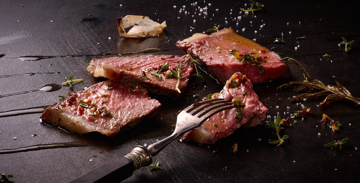 Dry Aged T-Bone Steak gegrillt in Scheiben geschnitten mit Gabel auf dunklem Metall Untergrundquerformat dark food photography münchen
