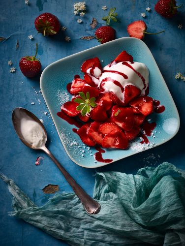 Desserts erdbeeren mit sauerrahmeiscreme himbeersauce auf teller aussenrum löffel tuch loose erdbeeren holunderblüten blauer untergrund gekleckse