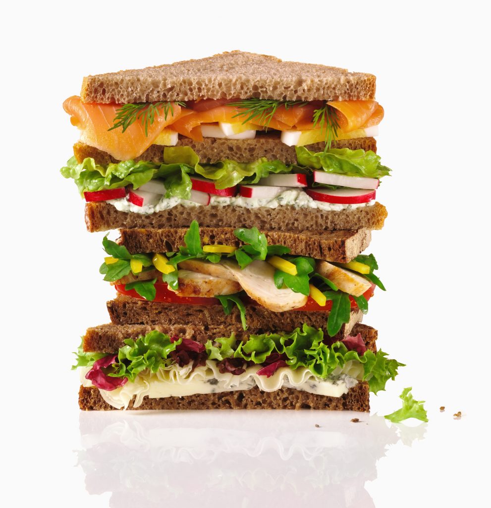 Food Allegorien ein riesengrosses Sandwich mit sehr vielen Schichten aus Salat Wurst Käse Lachs Gemüse türmt sich auf. weisser Hintergrund Freisteller