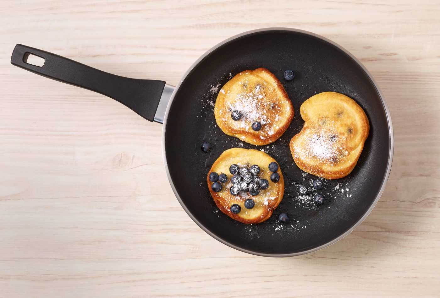 drei Blaubeerpfannkuchen werden in einer beschichteten Bratpfanne zubereitet. Sie sind mit Puderzucker bestreut. Food-Verpackungen