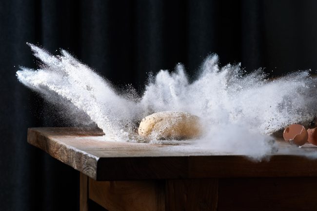 high speed fotografie von einem teigklumpen der in mehl faellt das mehl schiesst explosionsartig durch die luft auf altem holztisch grauer vorhang als hintergrund