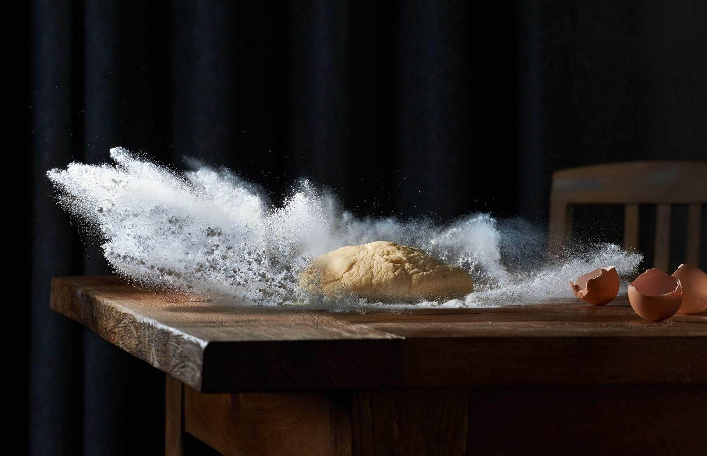 high speed fotografie von einem teigklumpen der in mehl faellt das mehl schiesst explosionsartig durch die luft auf altem holztisch grauer vorhang als hintergrund