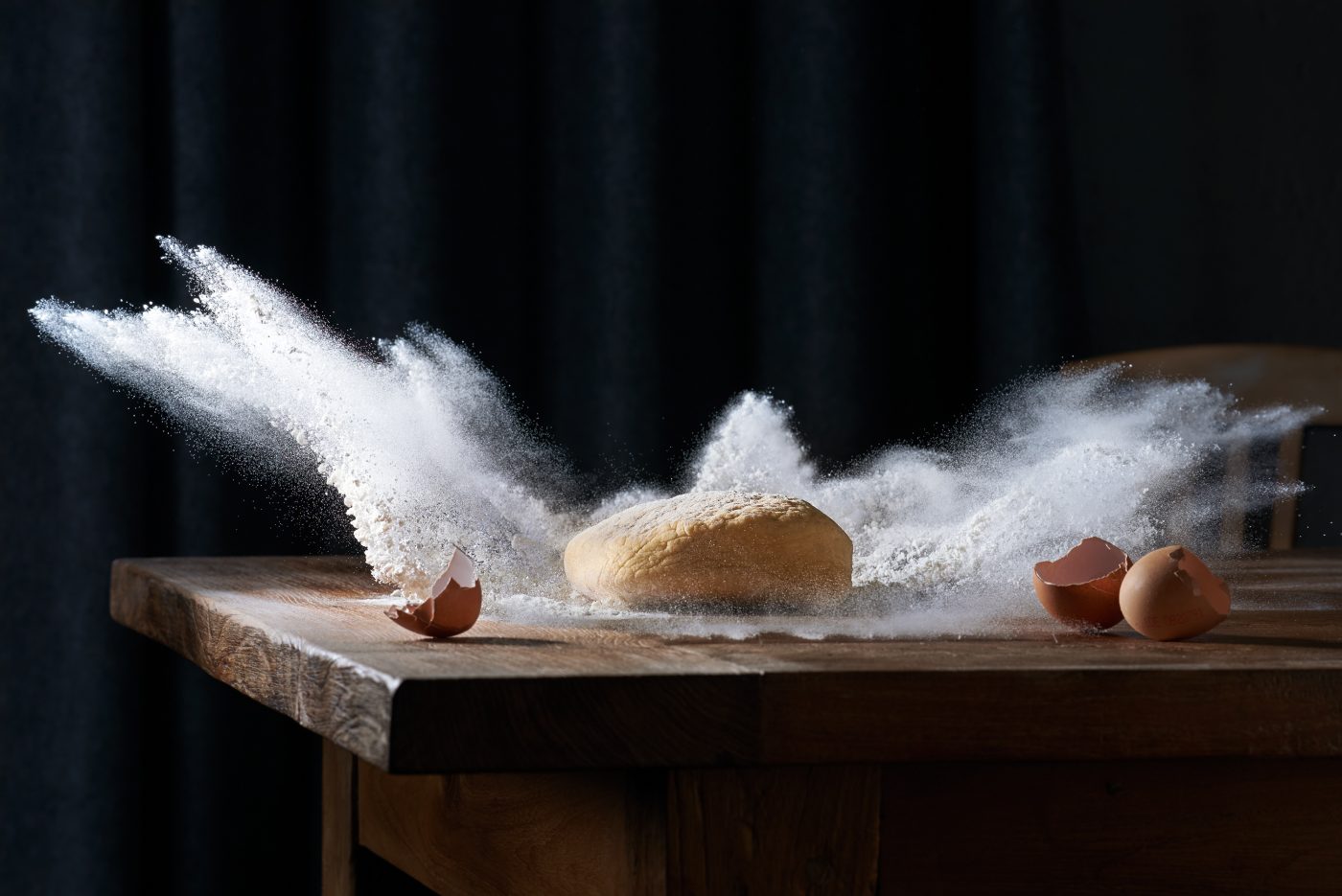 high speed fotografie von einem teigklumpen der in mehl faellt das mehl schiesst explosionsartig durch die luft auf altem holztisch grauer vorhang als hintergrund