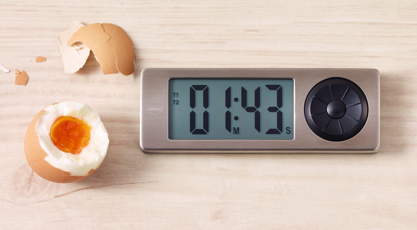 Food-Verpackungen Foto mit Eieruhr. Daneben liegt ein aufgebrochenes Ei mit Eierschale