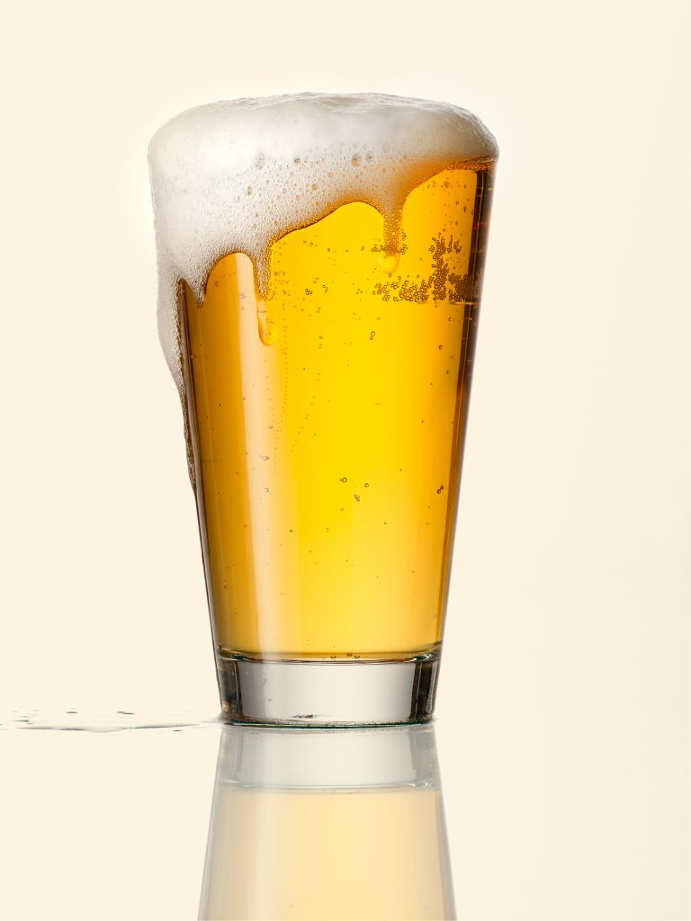 Bier ist in einem Glas randvoll eingeschenkt und läuft über Tautropfen auf dem Glas goldener Hintergrund