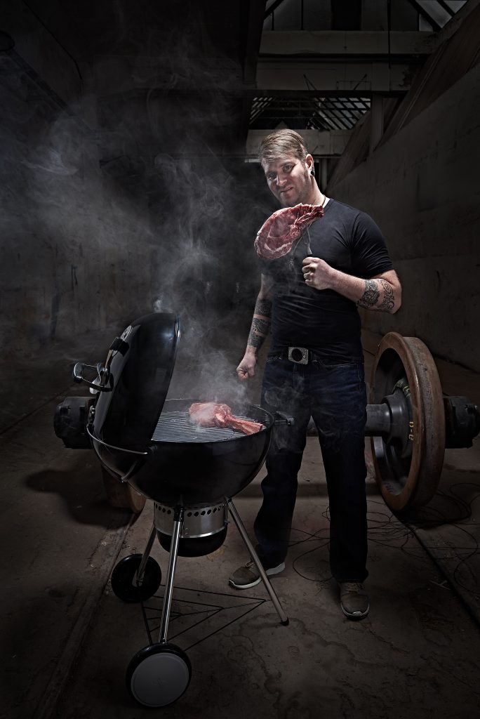 Man mit schrägem Gesichtsausdruck und Tattoos steht am Grill und hat eine Fleischgabel mit einem rohen Steak in der Hand. Dieser Hipster sieht cool aus. Rauch steigt aus dem Kugelgrill auf. Im Hintergrund sieht man eine alte Fabrikhalle von innen. Es ist im Hintergrund dunkel.
