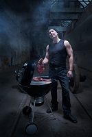 man with allot of muscles stands next to a barbecue, grilling meat in an industrial Hall, location shooting wih hipster model