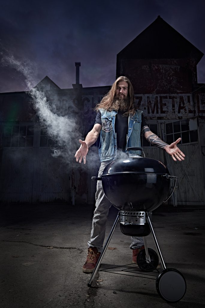 Man mit langen Haaren und Bart steht am Grill und breitet die Arme aus wie Jesus. Dieser Hipster sieht cool aus. Rauch steigt aus dem Kugelgrill auf. Im Hintergrund sieht man eine alte Fabrikhalle. Es ist Abenddämmerung und wird schon dunkel.