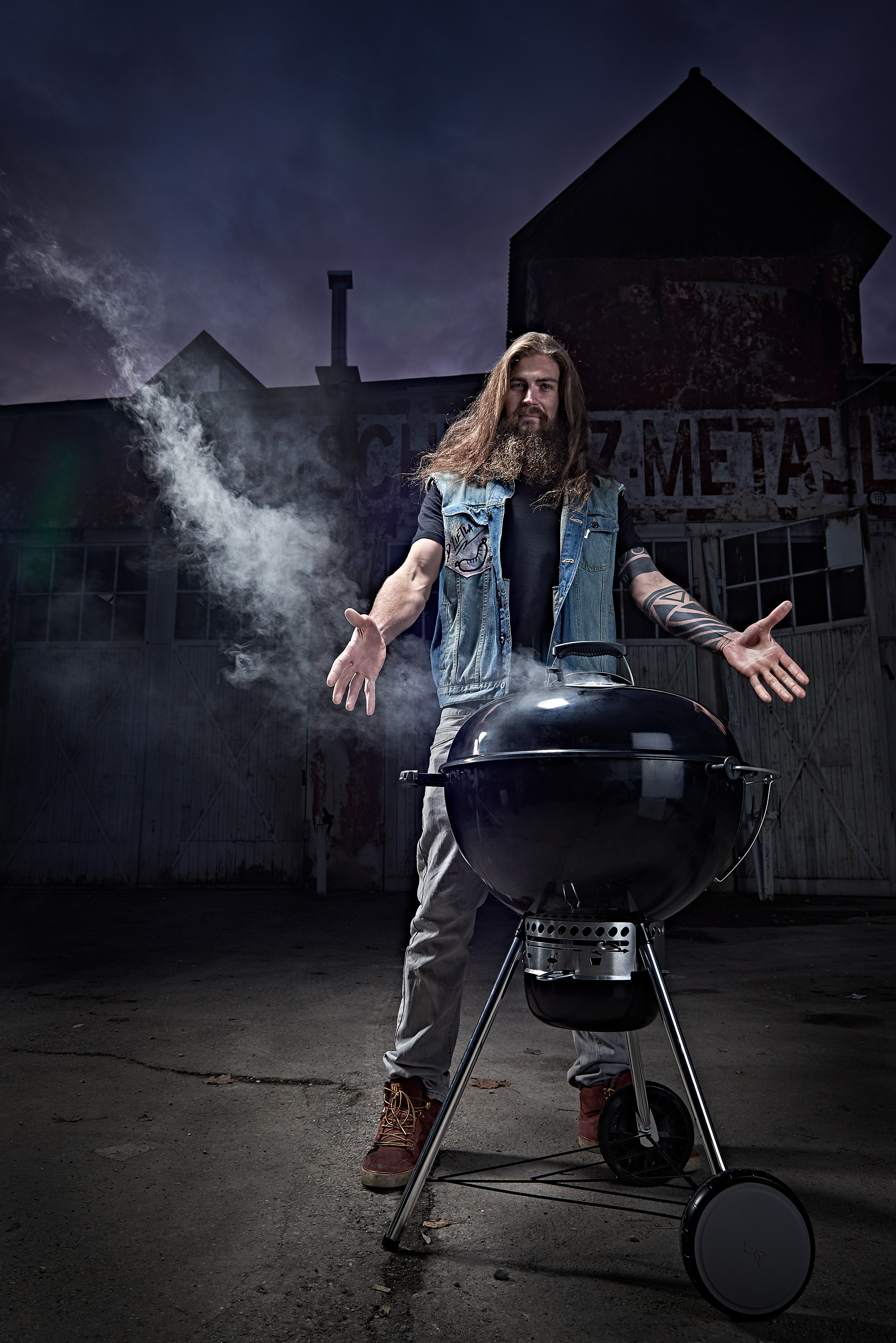 man with arms spread stands behind a barbecue, grilling dinner in front of a industrial location outside shooting