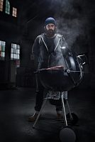 man with beard and tattoos stands behind a barbecue, grilling dinner in an industrial Hall, location shooting