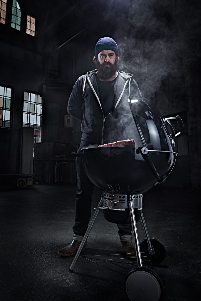 man mit bart und tattoos steht hinter einem grill auf dem ein saftiges rindfleisch liegt. er hat einen bart und mütze auf.