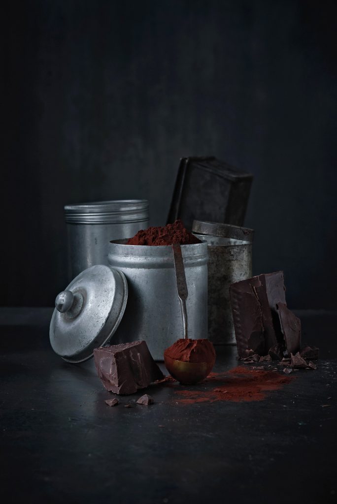 Zutaten zu einem Schokoladen Kuchen sind zu einer Gruppe arrangiert, Kakaopulver ist in einer Aludose, Schokoladenbrocken liegen auf dem dunklen Untergrund, mehrere ander Blechdosen und ein Metalllöffel mit Kakao