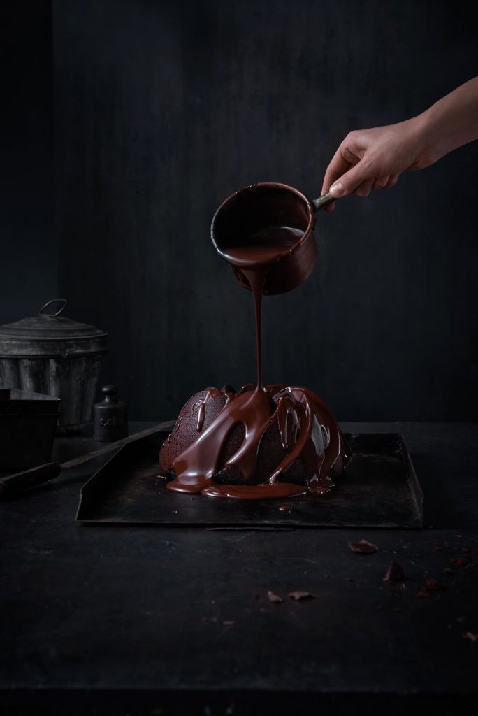 Schokoladen Kuchen wird mit warmer flüssiger Schokolade begossen schokoliert. Eine Hand hällt eine Kanne aus der die geschmolzene Schokolade fliesst. es liegen schokoladen bruchstücke rum. dunkler Untergrund dunkler Hintergrund