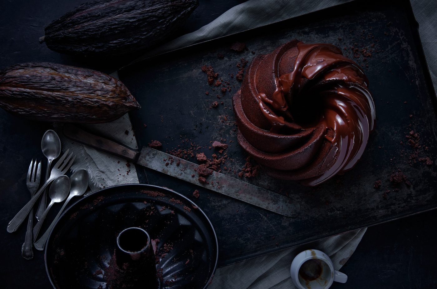 Schokoladen kuchen aus der Vogelperspektive fotografiert zwei ganze Kakaobohnen ein grosses Messer Besteck ausgetrunkene Espressotasse liegen aussen rum. sehr dunkle Stimmung, darkfoodphotography