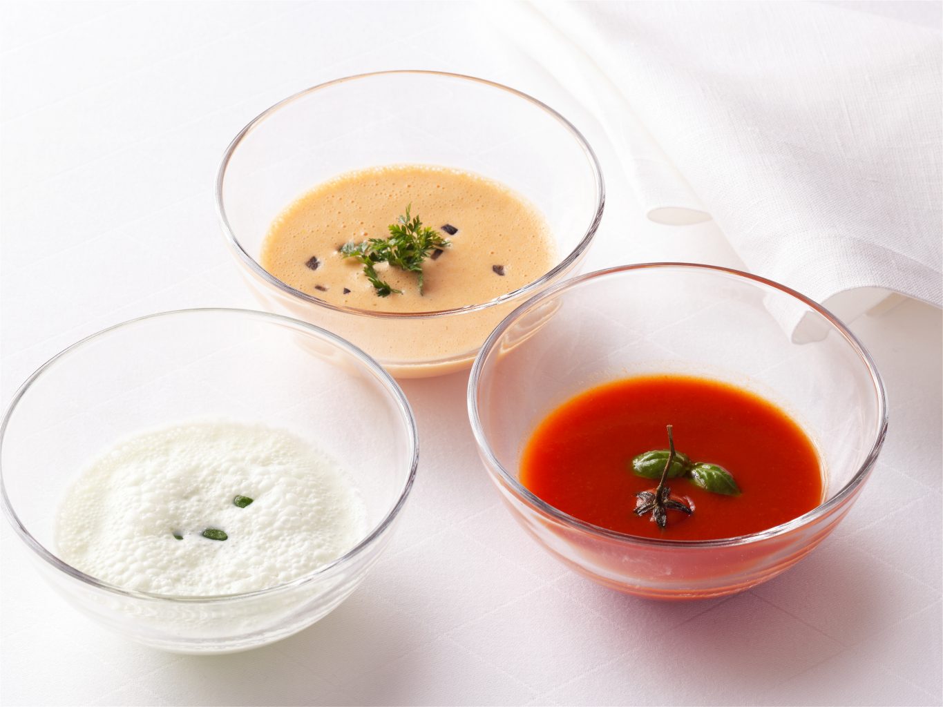 Verpackungsaufnahmen von drei Suppen in Glasschalen mit Kräutern auf weisser Tischdecke