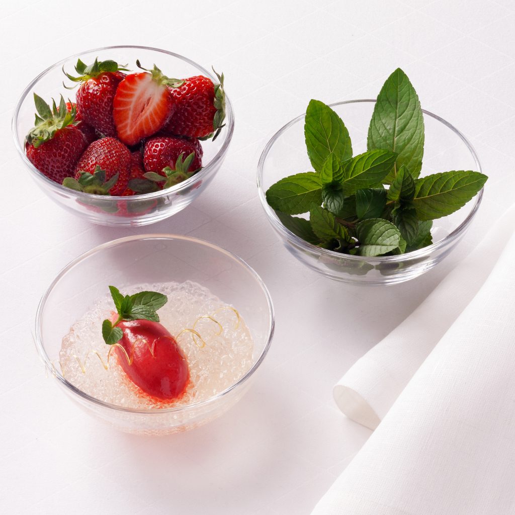 Verpackungsaufnahmen mit drei Glasschüsseln die mit Früchten, Minze und Nachspeisen gefüllt sind. Es steht auf einer weissen Tischdecke mit weisser Serviette.
