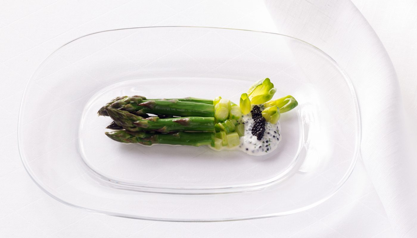 Verpackungsaufnahmen einer grossen Glasplatte mit Spargel, Kaviar und weisser Sauce. Der Teller steht auf einem Tisch mit weisser Tischdecke.