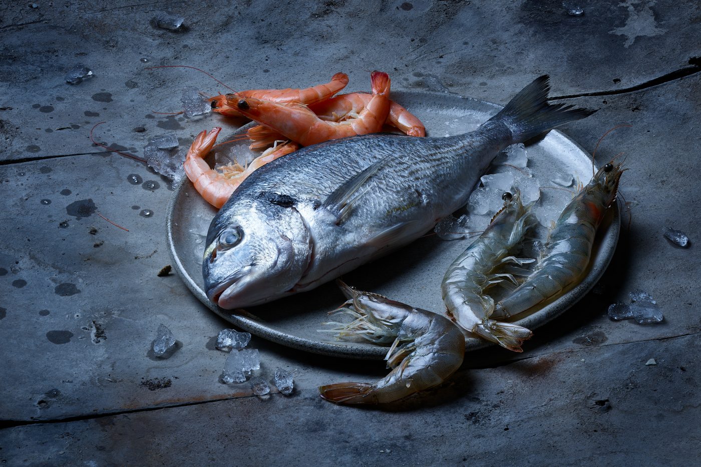 seafood bestehend aus einer ganzen rohen dorade mit rohen und gegarten krevetten. die fische liegen auf einem tonteller