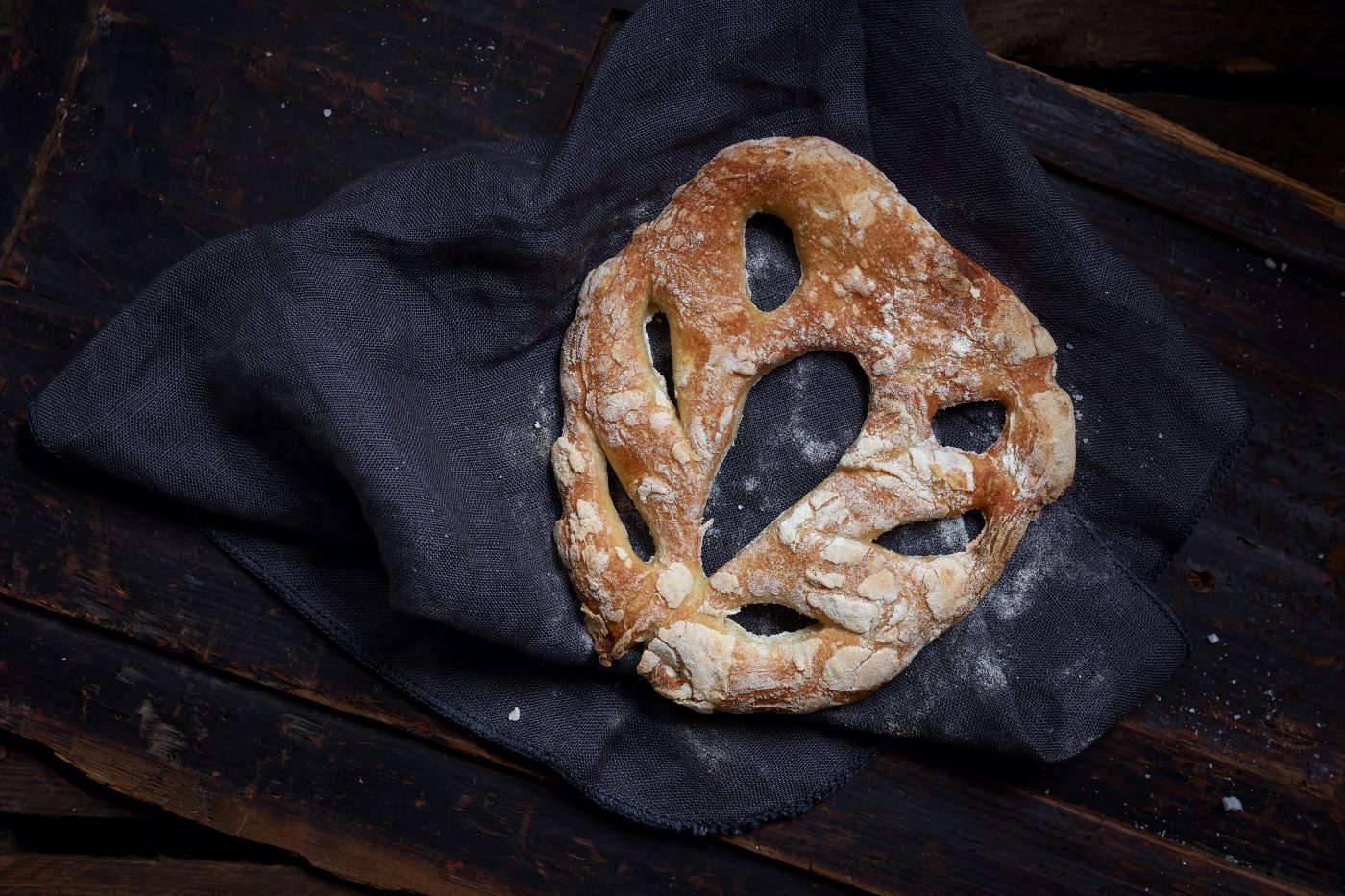 Brot Fougasse liegt auf dunklem Tuch. Das Set besteht aus dunklem Holz. Es liegen Krümmel im Set