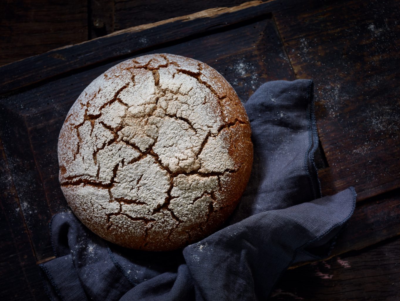 ein Laib Brot mit Rissen liegt auf dunklem Tuch. Das Set besteht aus dunklem Holz.