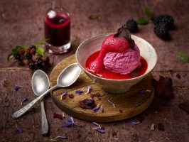 Eine wunderschöne Kugel Brombeereis liegt leicht angeschmolzen in einer handgetöpferten Schale. Sie ist mit einem Roto Bete Schaum garniert. In der Schale ist ausserdem Rote Bete Sauce. Die Schale steht auf einem uniquen Holzbrettchen. Zwei alte Silberlöffen liegen schön drapiert an den Bretchen. Im Umfeld sind Brombeerzweige mit reifen Brombeeren daran zu sehen. Ausserdem ist eine Glasfläschchen mit zusätzlicher Brombeersauce im Bild. Der Untergrund ist eine alte, lilane Holztafel. Nach hinten ist ein schönes Bokeh zu sehen. Die Perspektive ist leicht von oben. Fotografiert wurde dieses Bild mit einer Fuji GFX 100 Kamera bei Blende 8.