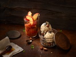 Zwei geriffelte Kugeln Haselnusseis liegen leicht angeschmolzen in einer alten Glasschale mit einem Metaldeckel der an der Schale anlehnt. Daneben steht eine weiteres Glas mit Pfirsichspalten, die in einer transparenten Vanillesauce eingedickt sind. Als Deko sind auch VAnillestangen im Glas. Auf dem rostigen Metaluntergrund liegen zerbrochene Haselnussstückchen, Mohn und grüne Kräuter. Zwischen beiden Gläsern lieht ein alter Silberlöffel. Im Vordergrund ist ein Honiglöffel, der mit Honig überzogen ist.  Der Hintergrund ist eine dunkle Holzwand. Nach hinten ist ein schönes Bokeh zu sehen. Die Perspektive ist leicht von oben. Fotografiert wurde dieses Bild mit einer Fuji GFX 100 Kamera bei Blende 8.