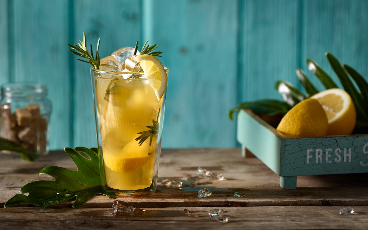 Tee im Glas mit Zitronen und Rosmarin. Aussen liegen in einem Instagram Set ein Tablet mit Zitronen. Frischer türkis Hintergrund aus Holz. Stylisch cooles Liquids Foto.