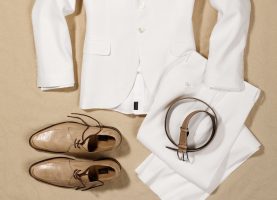 a light-coloured suit lies on the sand with shoes and belt as outfit, fashion still photography