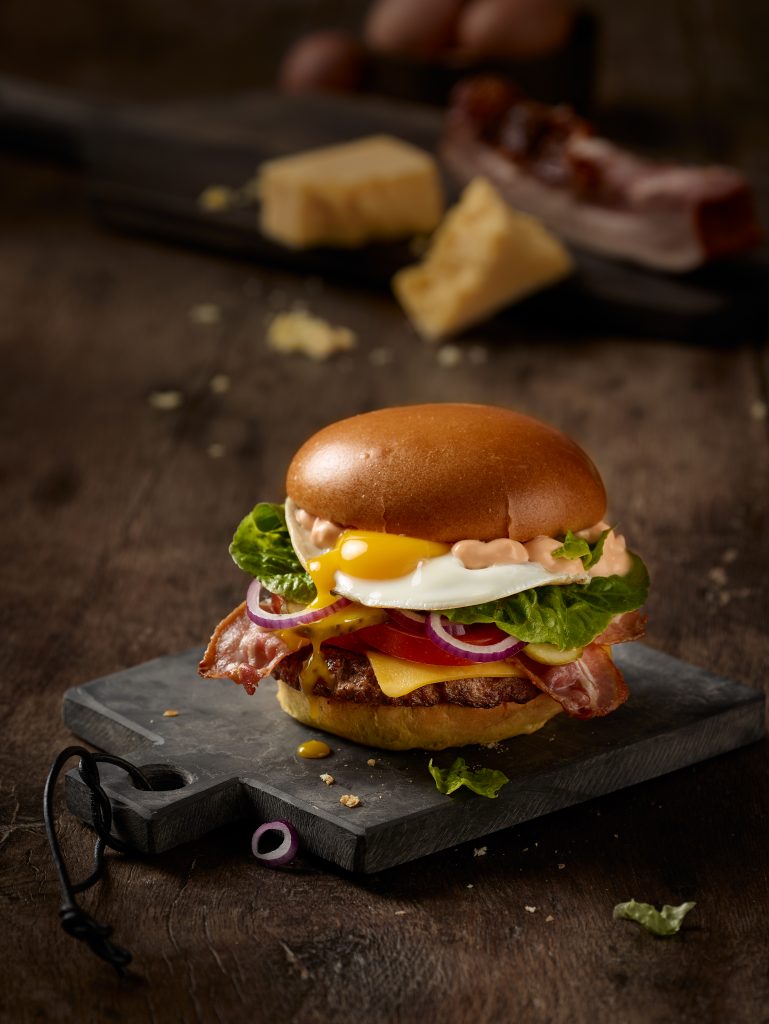 freshly prepared Classic Burger with a fried egg in the middle. Ingredients in th background.
