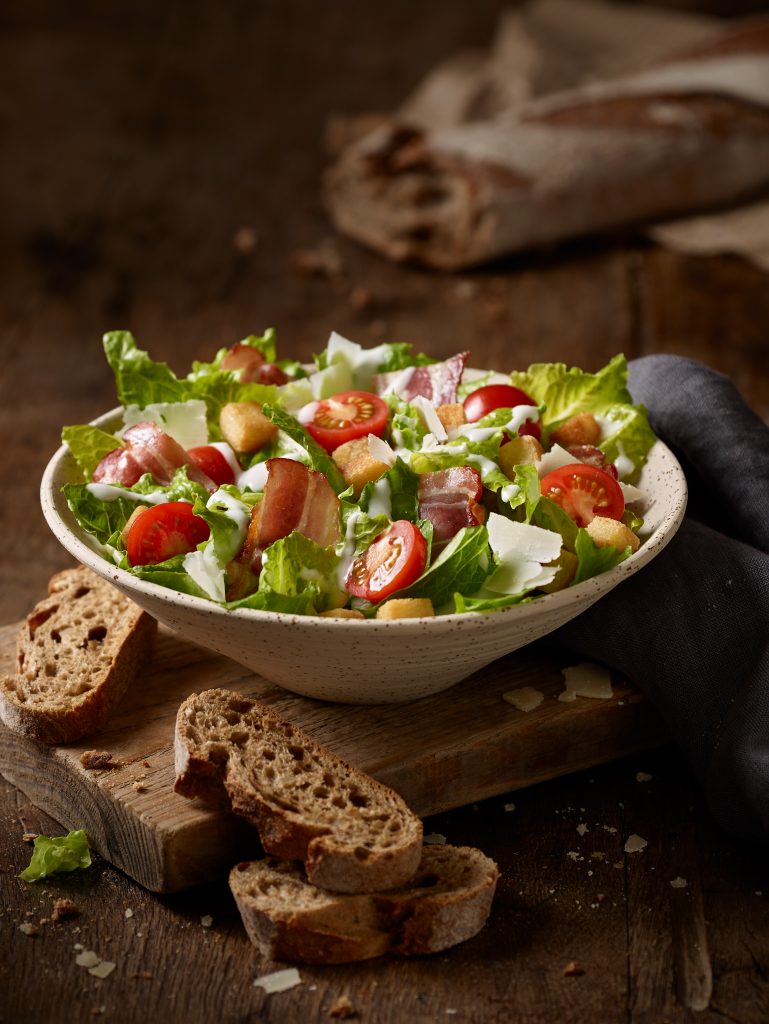 freshly prepared caesar salad in a bowl. The underground is made of wood, ingredients are in the background.