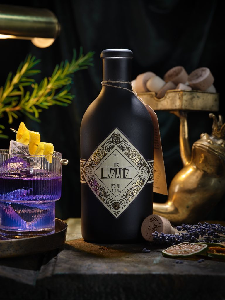 Wir sehen eine Flasche Ilusionist Gin mit Gintonic daneben. Es ist eine Zitronenzeste zu sehen. Ei Frosch hält viele Korken.