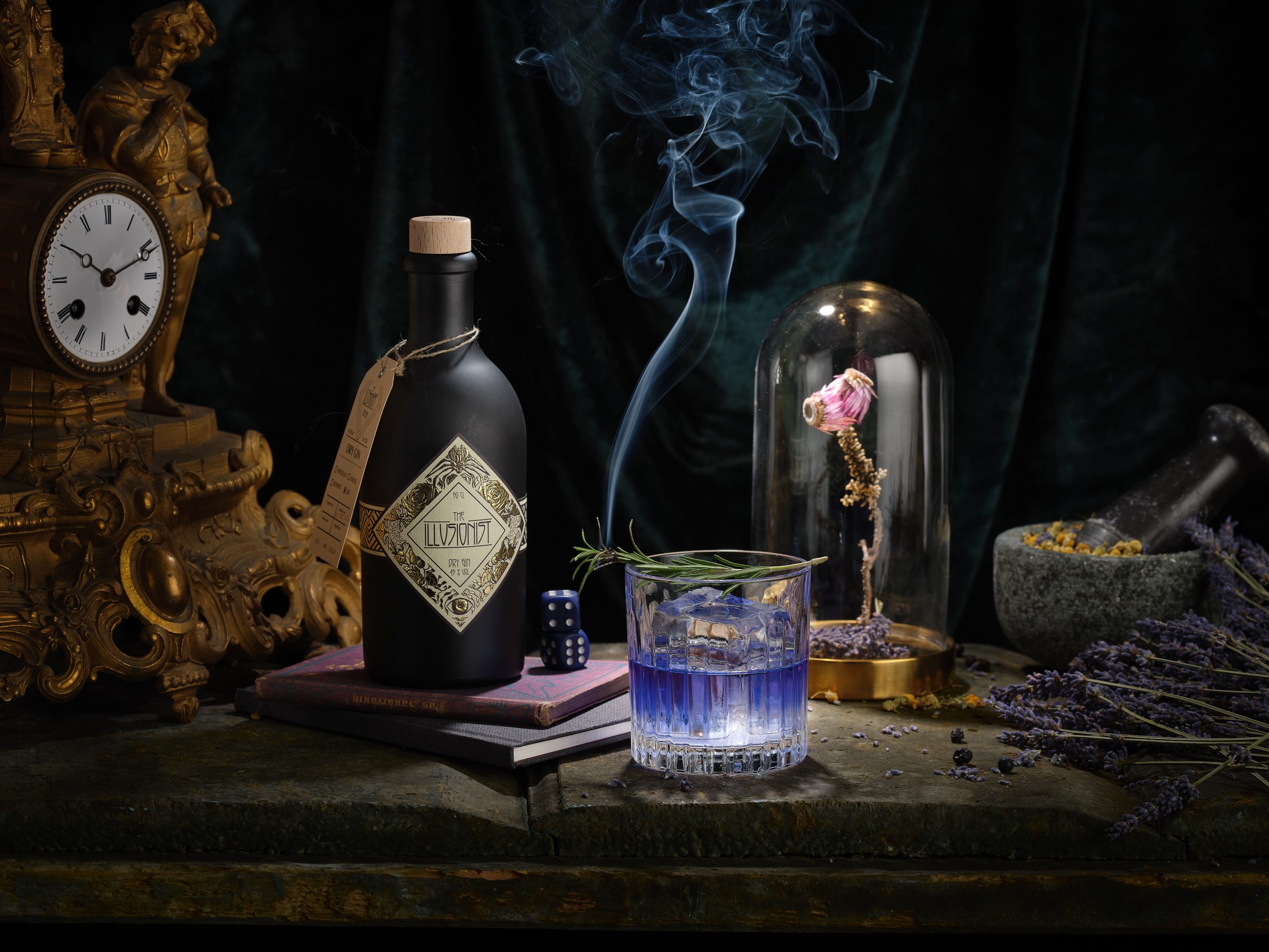 EIne Flasche Gin steht neben einem Gintonic mit Eiswürfeln. Auf dem Glas ist ein Rosmarinzweig angezündet und raucht. Neben dran steht eine alte Uhr und ein Glaskolben unter dem eine Blume zu sehen ist.