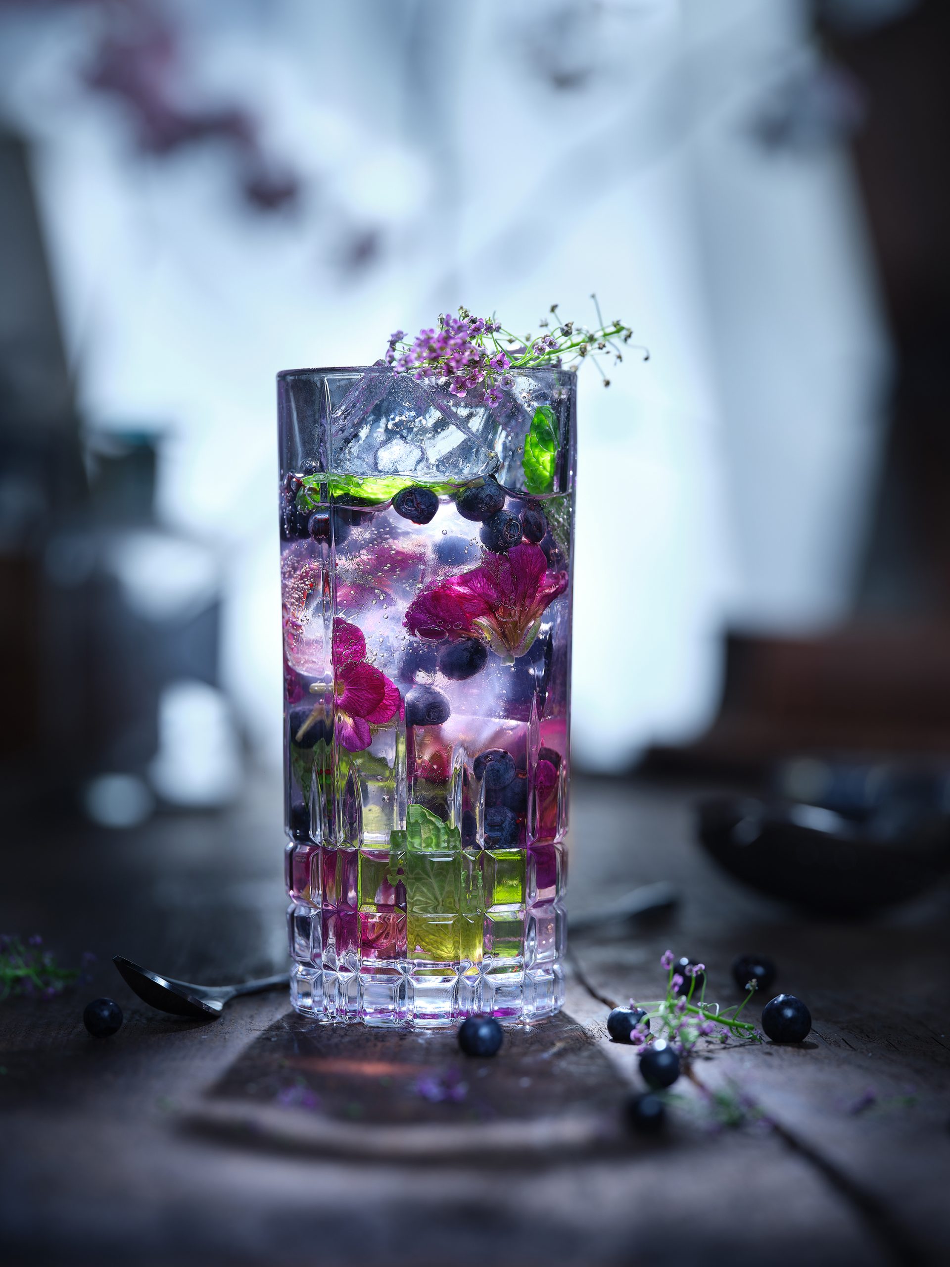 A blueberry gin and tonic cocktail stands in a bar environment. The background is very blurred.