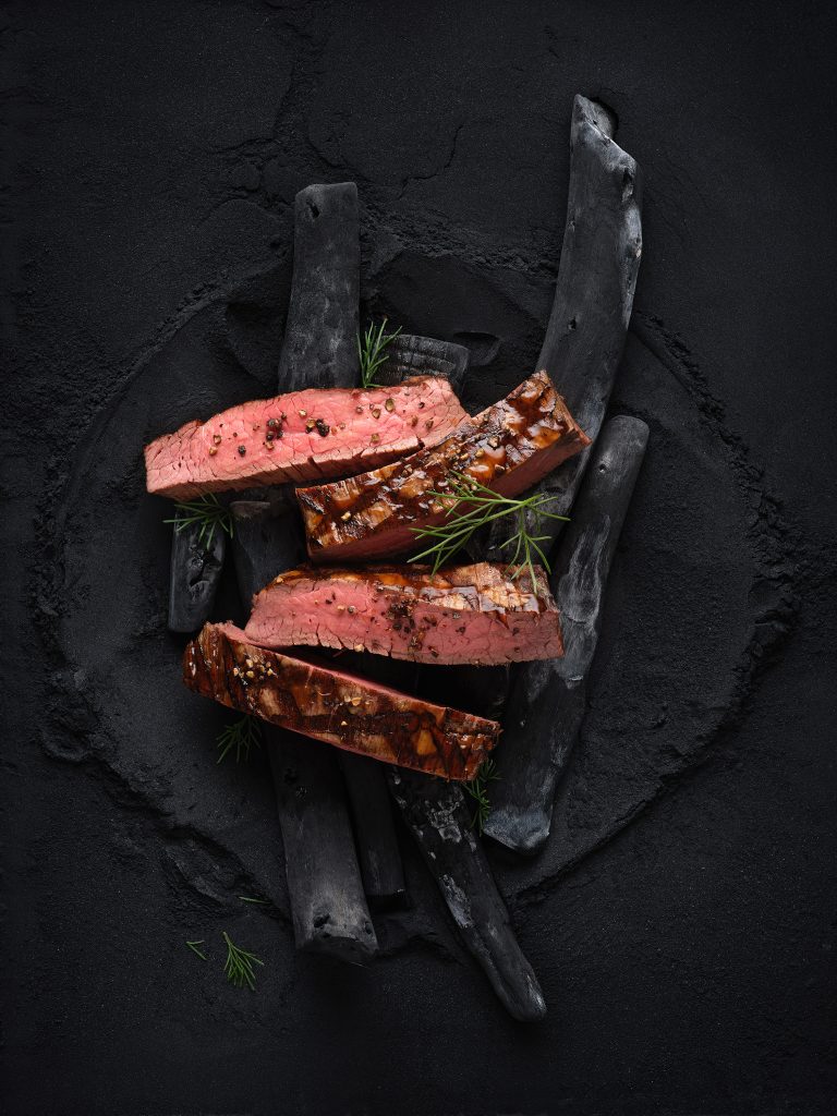 four sliced strips of flank steak lie on black charcoal. Everything lies on a dark black powder background