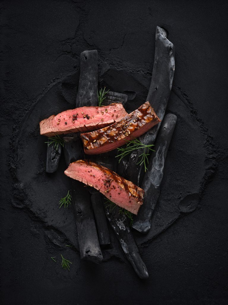 three sliced strips of flank steak lie on black charcoal. Everything lies on a dark black powder background